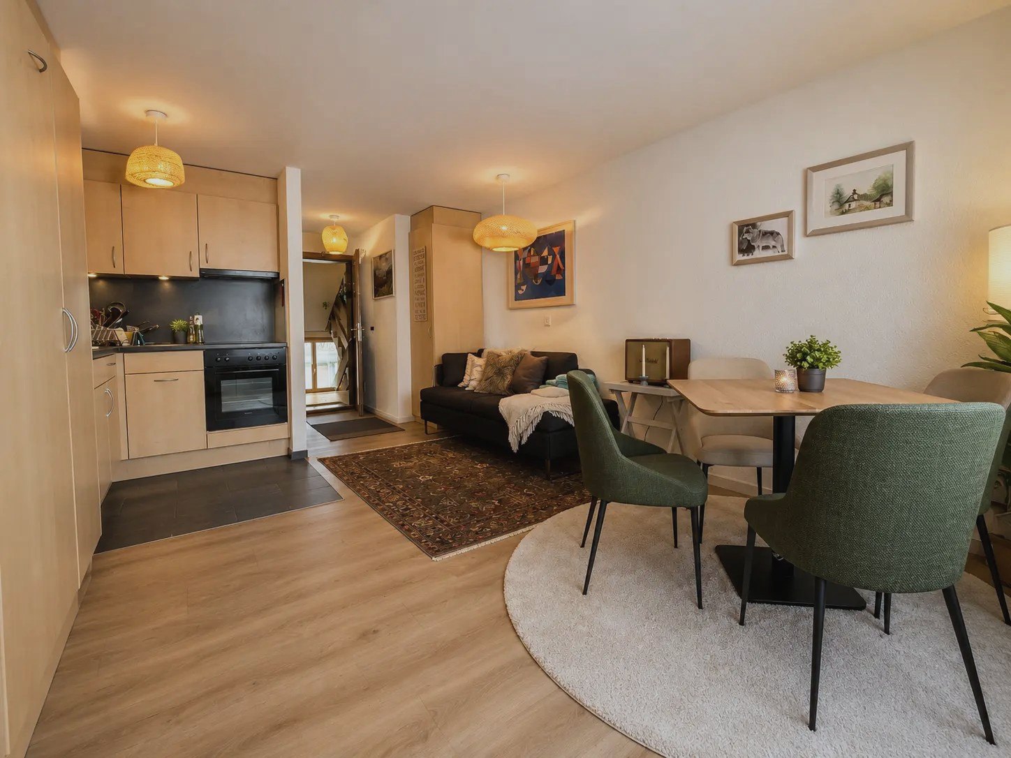 Essbereich des Studios Malo mit grünen Stühlen, rundem Teppich und Blick in die Küche
