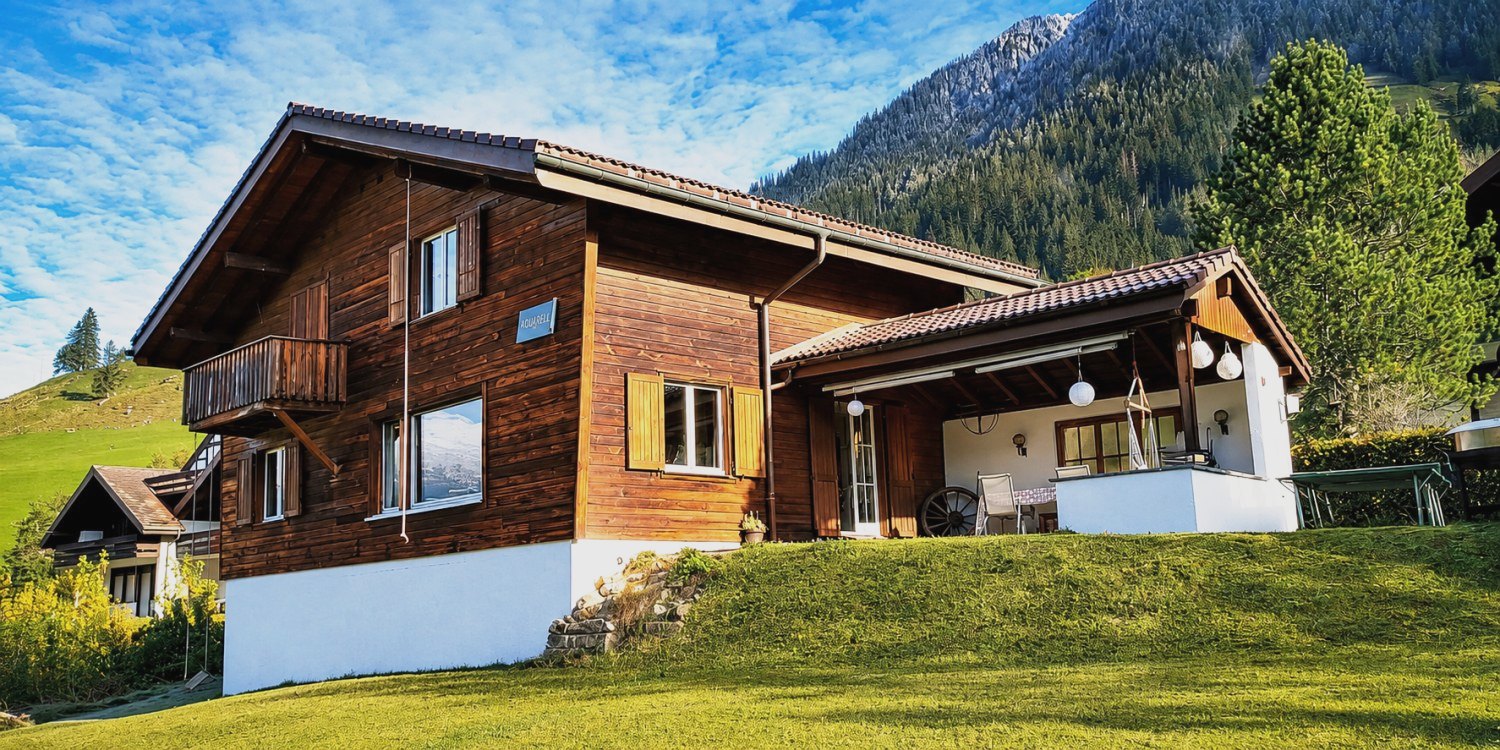 Chalet Nördli in Sörenberg: dunkles Holzchalet mit Balkon und Terrasse auf grüner Wiese vor Bergwald